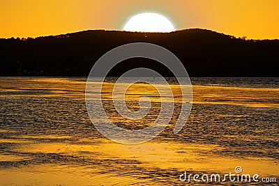 Sun glowing behind hill at lake by golden shimmering water and sky Stock Photo