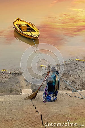 A yellow wooden boat float at sunrise close to a Indian lady dressed in blue, sweeping the stairs at the holy river Ganges Editorial Stock Photo