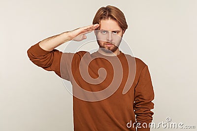 Yes sir! Portrait of patriotic guy with beard in sweatshirt saluting and listening to command, ready to obey Stock Photo