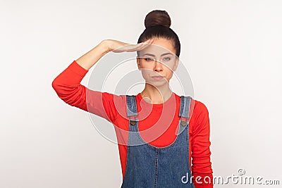 Yes sir! Portrait of strict responsible girl with hair bun in denim overalls giving salute listening to order with serious Stock Photo