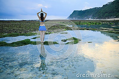 Yoga pose. Woman standing on the rock, practicing yoga. Young woman raising arms with namaste mudra at the beach. View from back. Stock Photo