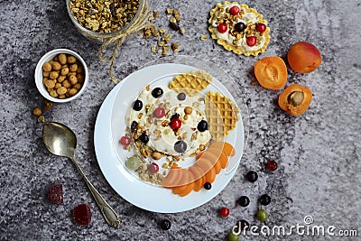 Yogurt with granola, berries and waffles on a white plate and nuts, peaches on a gray marble background. Healthy and Stock Photo