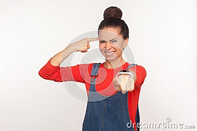 You are dumb, out of mind! Portrait of girl with hair bun in denim overalls showing stupid gesture and pointing to camera Stock Photo