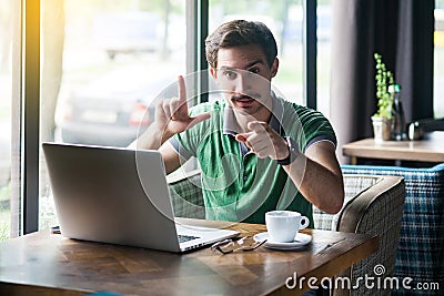 You are loser. Young serious businessman in green t-shirt sitting, pointing and showing loser gesture and looking at camera Stock Photo