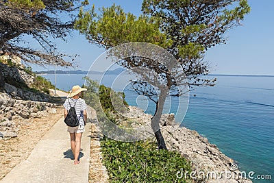 Young active feamle tourist wearing small backpack walking on coastal path among pine trees looking for remote cove to Stock Photo