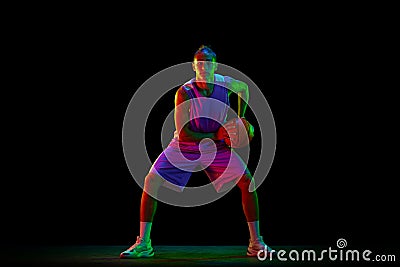 Young, active, motivated guy, basketball player in uniform standing in position with ball against black studio Stock Photo