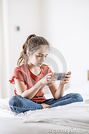 Young addicted gamer girl plays on a phone while sitting on a bed at home Stock Photo