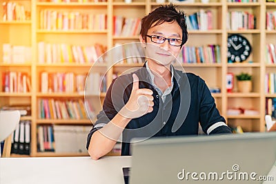 Young adult Asian man with laptop, thumbs up ok sign, home office or library scene, with copy space, success or technology concept Stock Photo