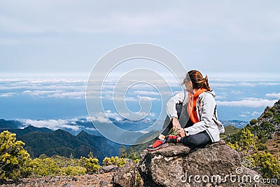Young adult caucasian female dressed sporty clothes resting on the rock and enjoying clouds view from Pico Ruivo mount 1861m - Stock Photo