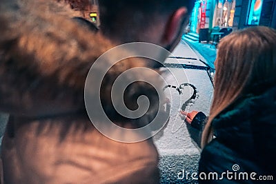 Young adult couple draws a heart on snow covered car Stock Photo