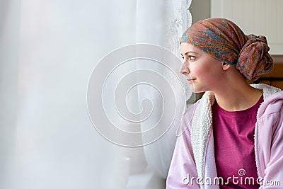 Young adult female cancer patient wearing headscarf and bathrobe sitting in the kitchen. Stock Photo