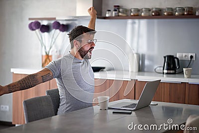 Young adult man with beard and tattoo yawns, stretching in front of laptop on table with cup of coffee and cell phone. lifestyle Stock Photo