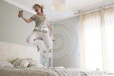 Young adult pretty woman go crazy for happiness and jump on the bed with coffee cup - overjoyed female caucasian people alone at Stock Photo