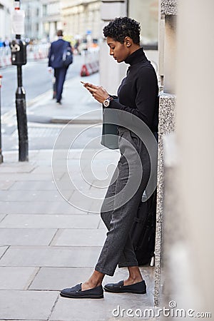 Young adult woman leaning on a wall in a London street using her smartphone, full length Stock Photo