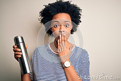Young African American afro tourist woman with curly hair holding thermo with water cover mouth with hand shocked with shame for Stock Photo
