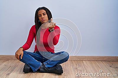 Young african american with braids sitting on the floor at home showing middle finger, impolite and rude fuck off expression Stock Photo