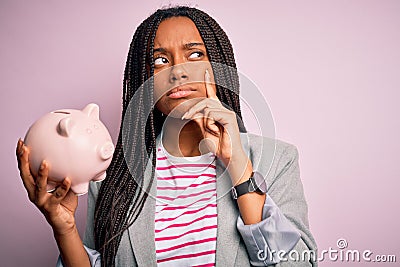 Young african american business woman saving money on piggy bank over background serious face thinking about question, Stock Photo