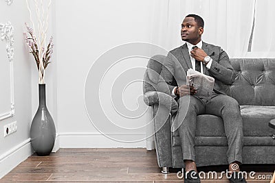 Young African American businessman in a gray suit reading a newspaper while sitting on a sofa. Stock Photo