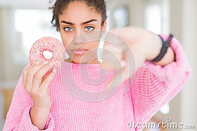 Young african american girl eating sweet pink donut with angry face, negative sign showing dislike with thumbs down, rejection Stock Photo