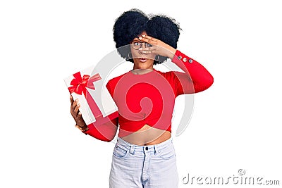 Young african american girl holding gift peeking in shock covering face and eyes with hand, looking through fingers with Stock Photo