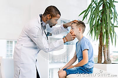 Young African American male doctor examines the eyes of his patient little boy, sitting on the couch in medical office Stock Photo