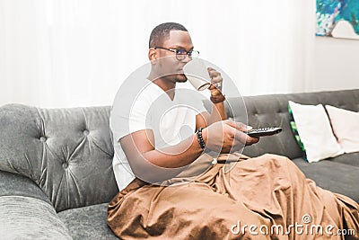 Young African American man, covered with a blanket, watching TV at home. Stock Photo