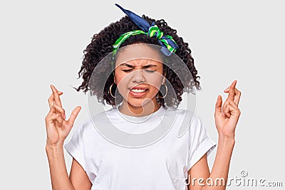 Young African American woman clenches teeth, raises fingers crossed, makes desirable wish, wears casual white t-shirt. Wishful Stock Photo