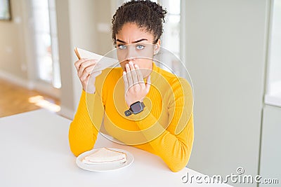 Young african american woman eating a sandwich as healthy snack cover mouth with hand shocked with shame for mistake, expression Stock Photo