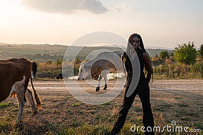 Young african girl in black clothes stands among the field, holds her hands behind her back, cows graze on the Stock Photo