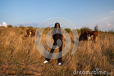 Young african girl in black clothes stands among the field, holds her hands behind her back, cows graze on the Stock Photo