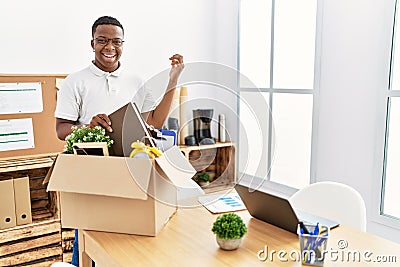 Young african man putting office objects into cardboard box celebrating victory with happy smile and winner expression with raised Stock Photo