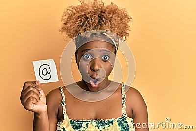 Young african woman with afro hair holding online mail symbol on paper scared and amazed with open mouth for surprise, disbelief Stock Photo