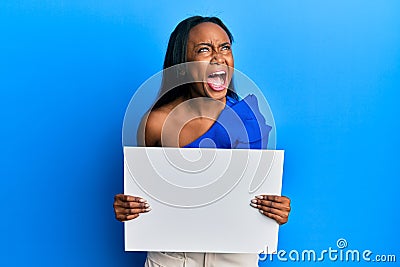 Young african woman holding blank empty banner angry and mad screaming frustrated and furious, shouting with anger looking up Stock Photo