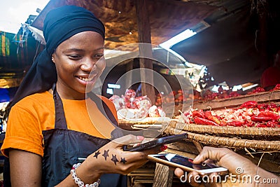 Young black woman selling tomatoes in a local african market receiving payment via mobile phone transfer Stock Photo