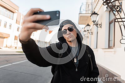 Young American hipster man in fashionable hoodie hooded in sunglasses in a stylish cap with a backpack is standing on the street Stock Photo