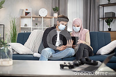 Young arabian couple in medical masks, feeling sick, sitting at home for isolation during covid-19 pandemic, and reading Stock Photo