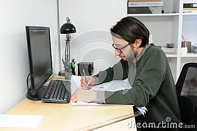 Young architect male In casual wear working in office on desk, types on a laptop, blueprints laying on her desk Stock Photo