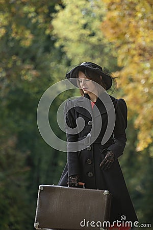 Young aristocratic woman in black coat and wide-brimmed hat with retro bag on autumn trees background. Woman in vintage clothes Stock Photo
