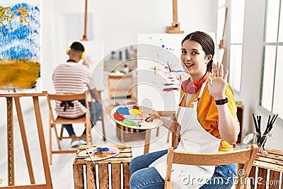 Young artist woman painting on canvas at art studio waiving saying hello happy and smiling, friendly welcome gesture Stock Photo