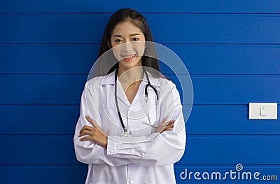Young asian doctor in white gown and stethoscope stand with arm cross in front of blue wall Stock Photo