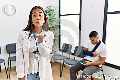 Young asian doctor woman at waiting room with a man with a broken arm looking at the camera blowing a kiss with hand on air being Stock Photo