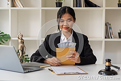 Young Asian lawyer holding a money envelope with a hammer and a laptop goddess of injustice on the table to the concept of corrupt Stock Photo