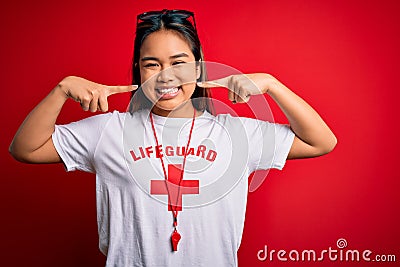 Young asian lifeguard girl wearing t-shirt with red cross using whistle over isolated background smiling cheerful showing and Editorial Stock Photo
