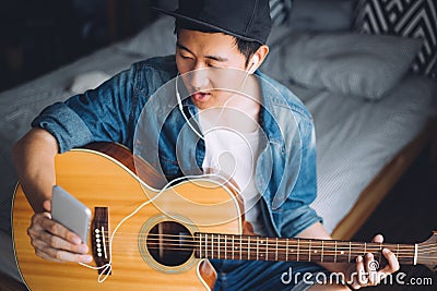 Young Asian man using a mobile phone with headphones while playing guitar in cozy bedroom. Stock Photo