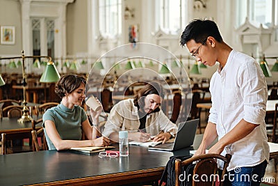 Young asian student in eyeglasses with backpack coming to study with friends in library of university Stock Photo