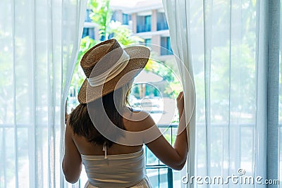 Young Asian traveler woman with straw hat looking outside the window in hotel room after check-in. Travel and summer holiday conce Stock Photo