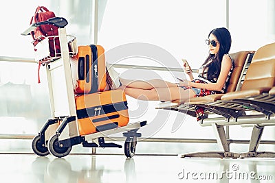 Young Asian traveler woman, university student sit using smartphone at airport, luggage and bag on trolley cart Stock Photo