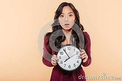 Young asian woman with curly long hair holding clock showing nearly 8 being late or missing something over peach background Stock Photo