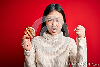 Young asian woman eating sweet and tasty belgian waffle over red isolated background annoyed and frustrated shouting with anger, Editorial Stock Photo