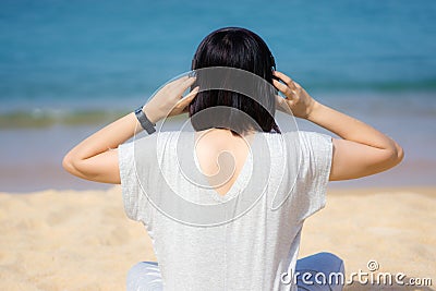 Young asian women in a gray dress wearing headphones, listening to music at the beach. Blue sky and crystal sea of tropical beach Stock Photo